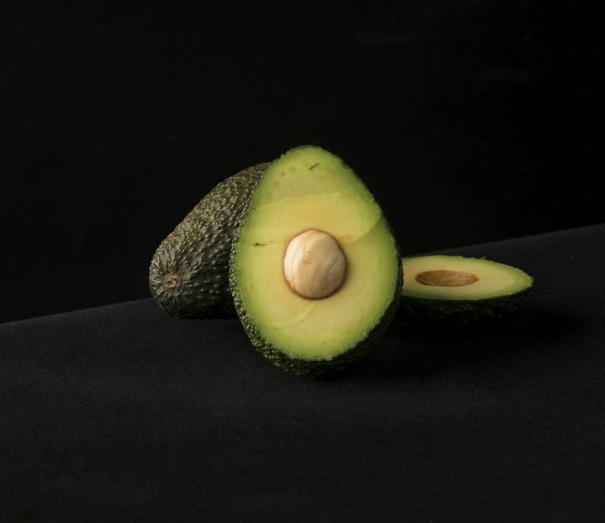 A halved avocado and a single slice on a near-black background, lit as a studio still life