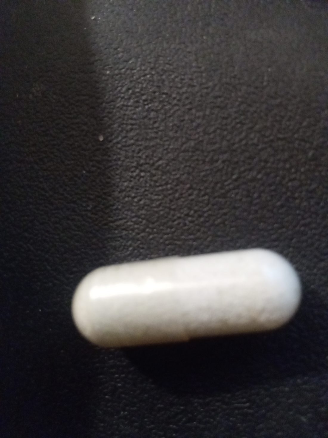 A single white capsule photographed on a dark textured surface