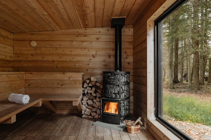 Interior of a wooden sauna with a lit wood burning stove, a stack of split logs, a bench with a rolled towel and a large window onto forest