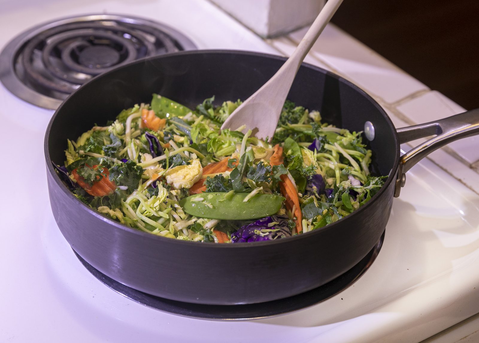 An overhead view of a large pan of colorful stir fried vegetables, including shredded kale, purple cabbage, broccoli, carrots, and snow peas, steaming on a stovetop with a wooden spoon resting in it
